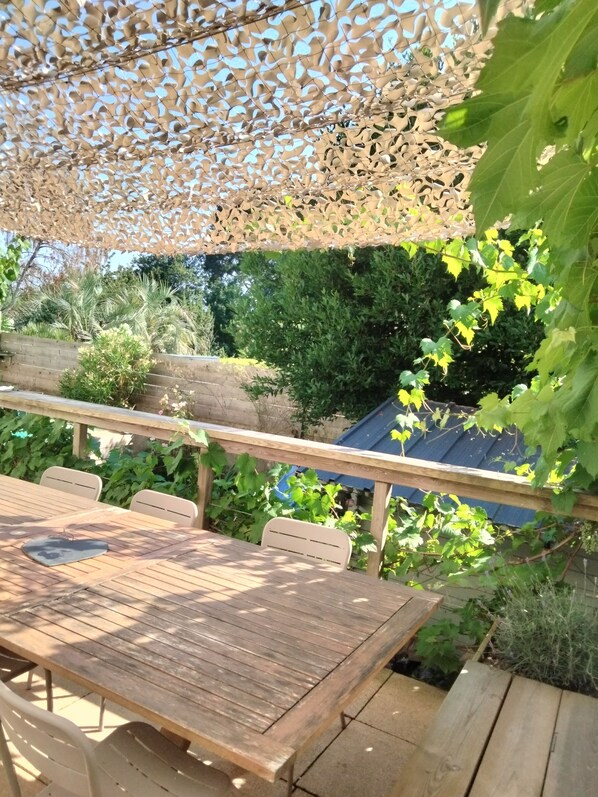 An outdoor patio with a wooden table and chairs, surrounded by greenery and a decorative lattice overhead.