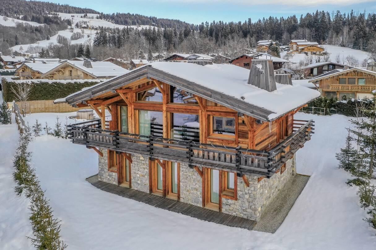 A log cabin with a balcony, stone foundation, and a snowy landscape in the background.