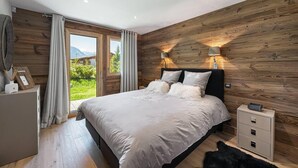 Un dormitorio con pared de madera, una cama con ropa de cama blanca, una mesita de noche y vistas a zonas verdes al exterior.