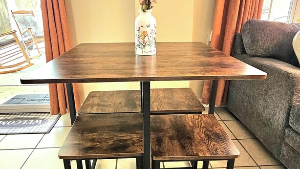 Una mesa de comedor de madera con patas metálicas, un jarrón blanco con flores, un cartel con la palabra 'welcome' y un sofá gris.