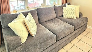 Una sala de estar con un sofá gris, un ventanal grande y piso de baldosas.