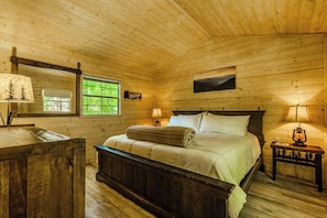 Een houten slaapkamer met een bed, nachtlampjes, een dressoir en een raam met uitzicht op groen.