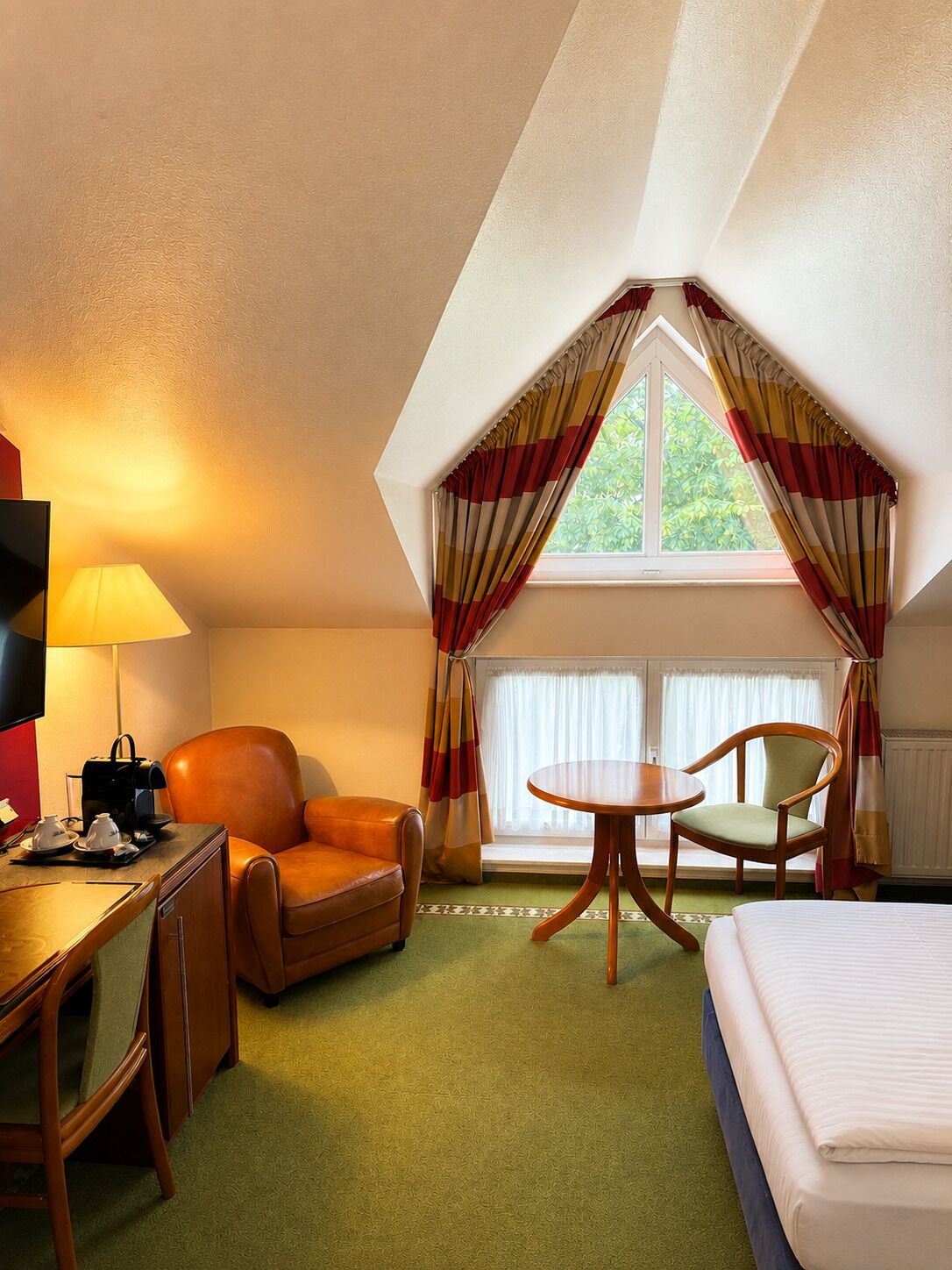 A hotel room with a bed, a desk with a lamp, a chair, a small table, and a window with curtains.