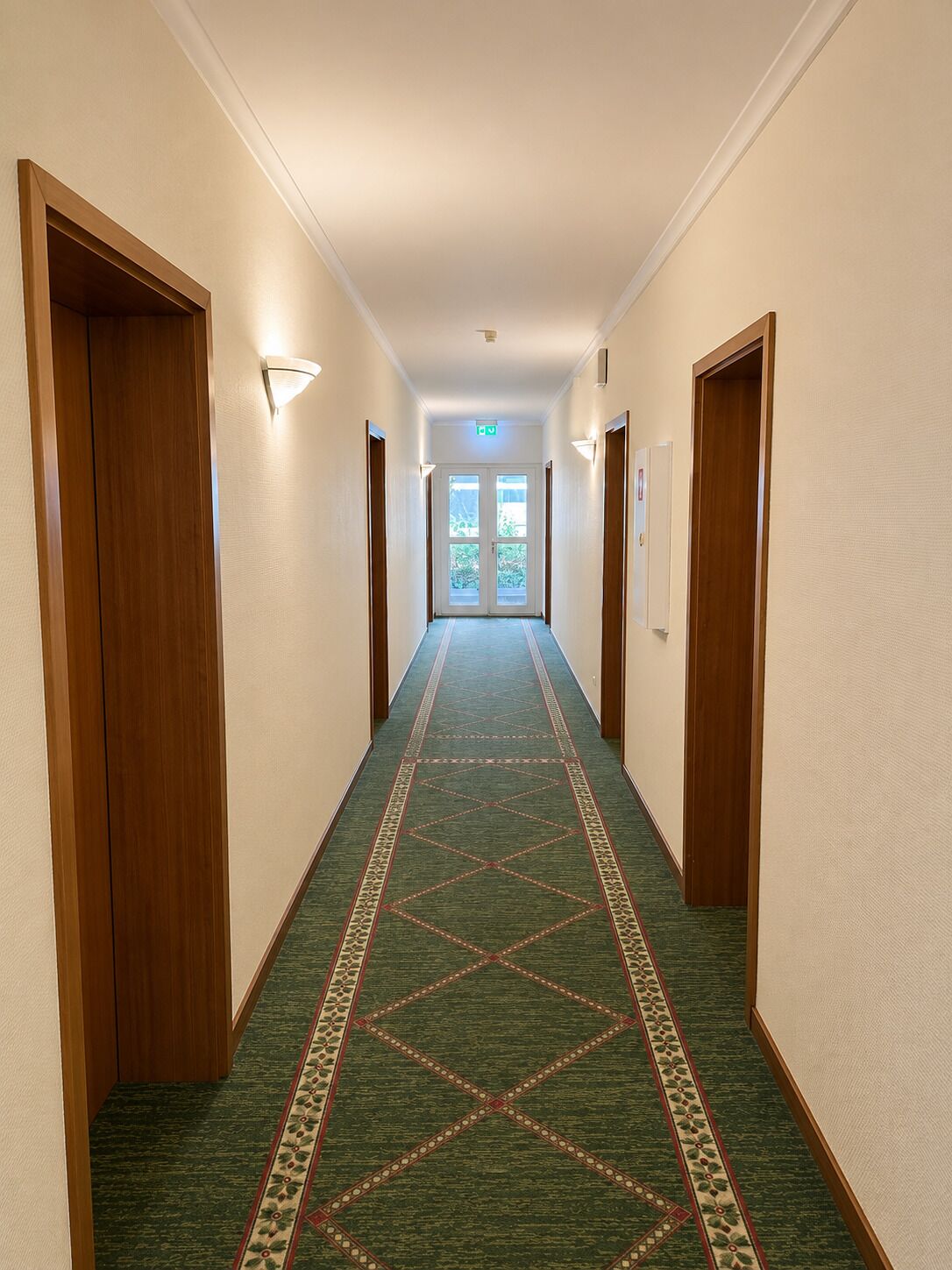 A hotel hallway with carpeted floors, wooden doors, and wall-mounted light fixtures.