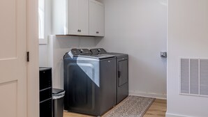 A compact laundry room with a front-loading washer and dryer, white cabinets above, and a patterned rug on the floor. - Chic Downtown Cody Hideaway (Cody)