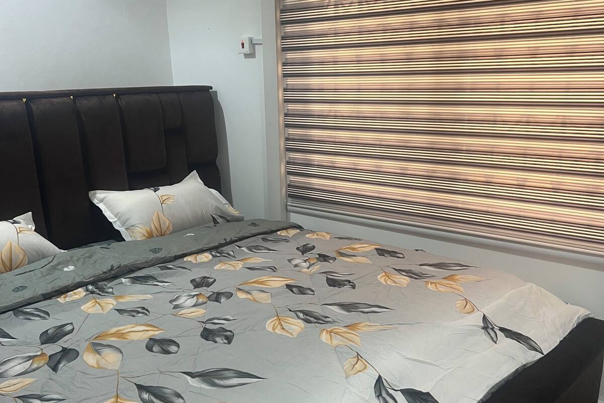 A bedroom with a bed, a headboard, a window with blinds, and an air conditioning unit.