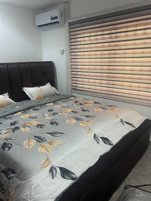 Un dormitorio con cama, cabecera, ventana con persianas y unidad de aire acondicionado.