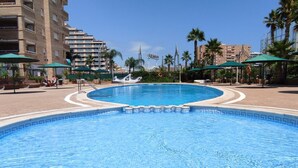 Piscina com borda em azulejo, cercada por palmeiras e guarda-sóis, com edifícios ao fundo.