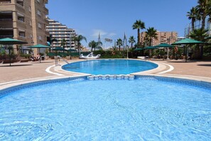 A swimming pool with a tiled edge, surrounded by palm trees and umbrellas, with buildings in the background. - ACV - Aguamarina-2ª linea planta 4 sur (OROPESA)