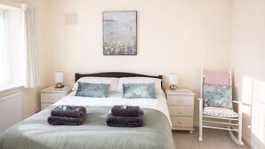 A neatly made bed with two pillows, a bedside table with a lamp, a chair with a floral cushion, and a painting on the wall.