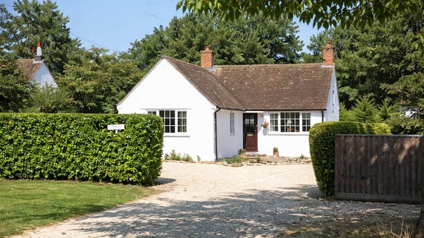 A white cottage with a gravel driveway, surrounded by greenery and a wooden fence.