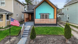 A modern house with a unique turquoise facade, a wooden porch, and a flag in the front yard. - New Indy Game House- Theater + Arcade - Sleeps 12 (Indianapolis)