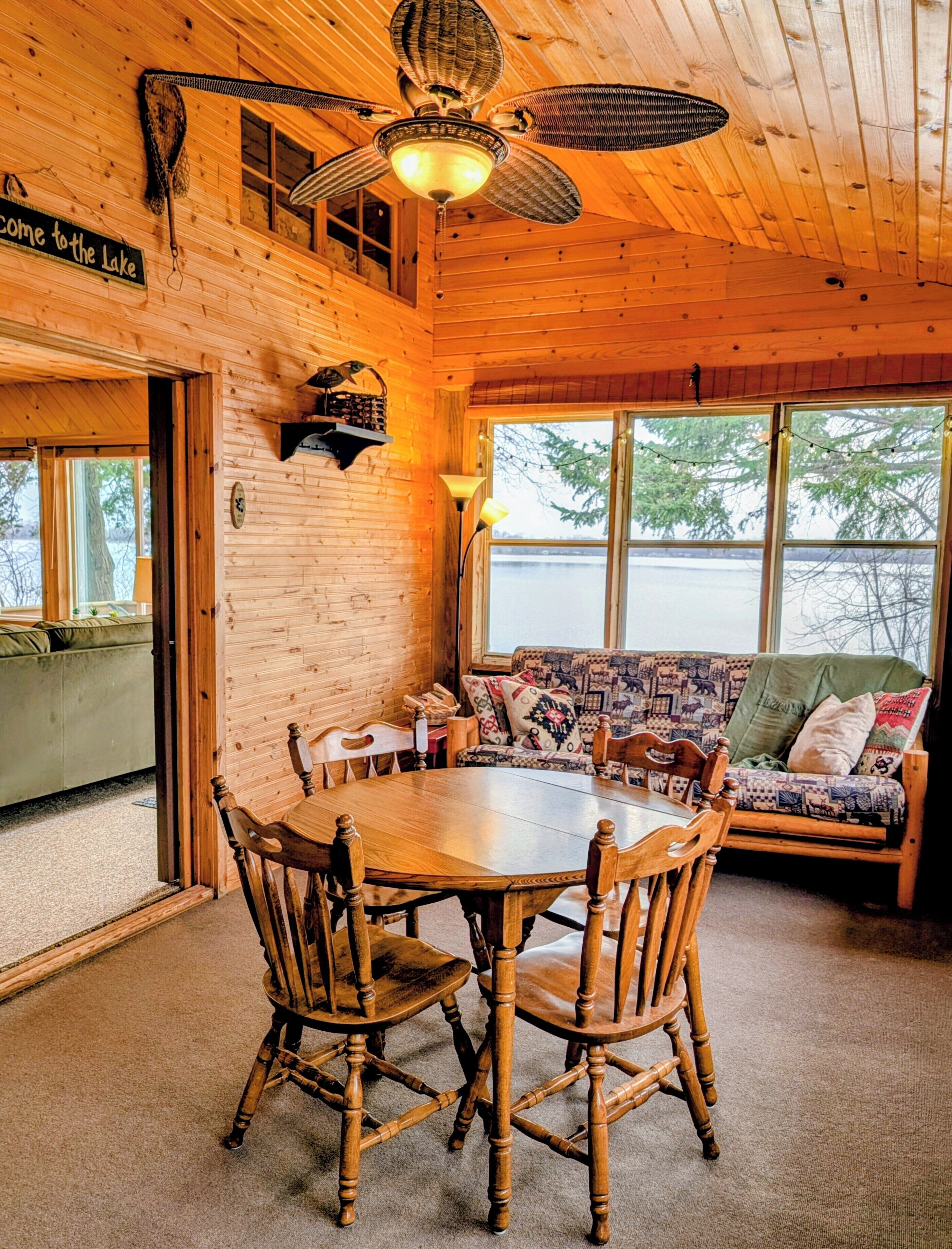 A wooden cabin interior with a dining table, chairs, a sofa, and a ceiling fan.