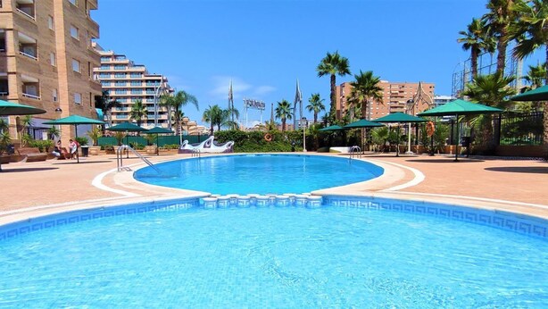 Ein Schwimmbecken mit einer gefliesten Kante, umgeben von Liegestühlen und Sonnenschirmen, mit einem Hotel im Hintergrund.