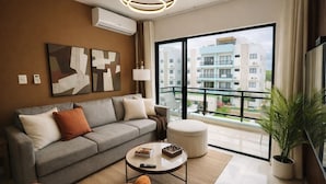 Un salon moderne avec un canapé gris, une table basse ronde, une plante en pot et une grande fenêtre donnant sur des immeubles.