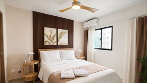Une chambre d’hôtel comprenant un lit, deux tables de chevet, un ventilateur de plafond, un appareil de climatisation, une fenêtre avec des rideaux et une œuvre d’art accrochée au mur.