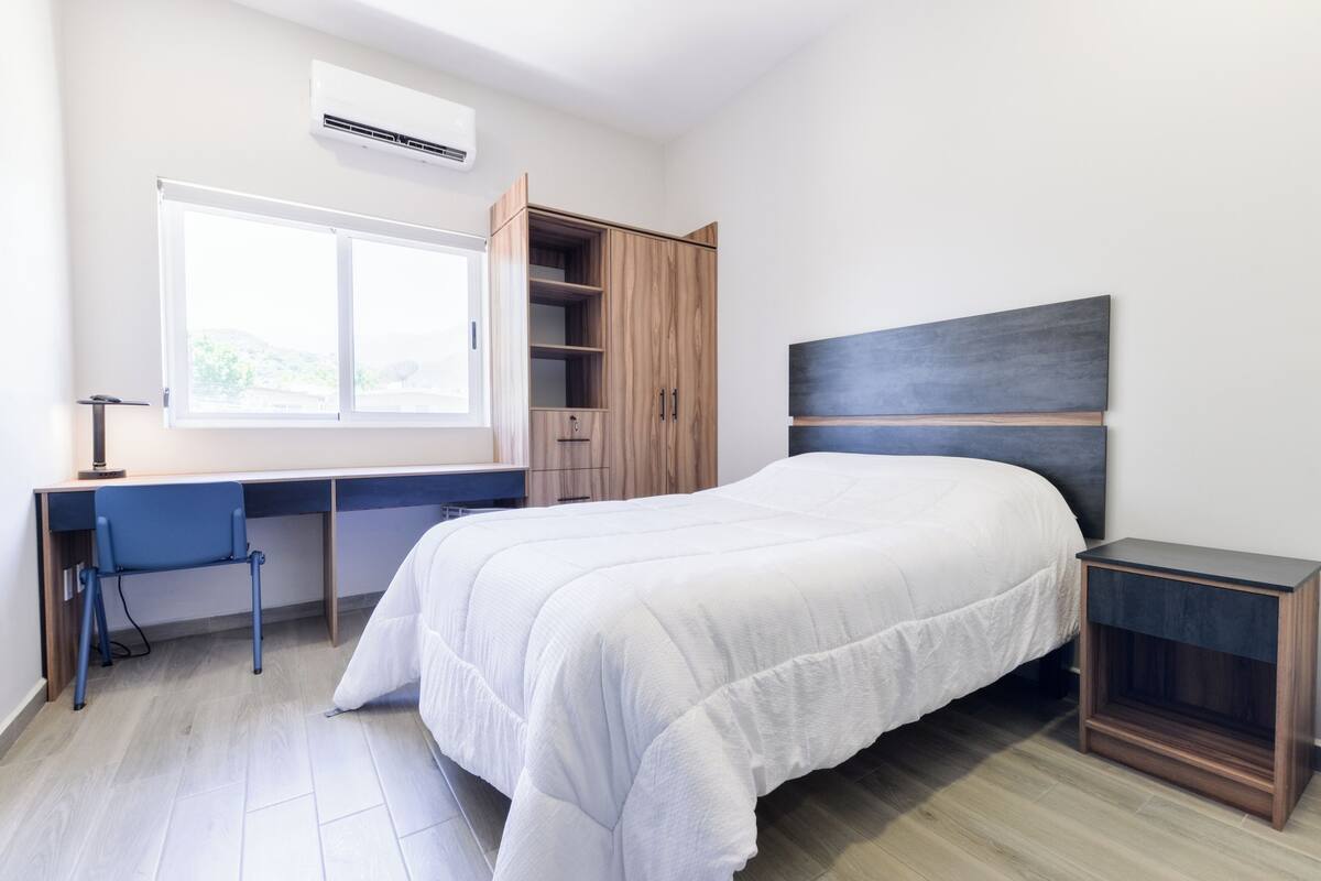 A bedroom with a bed, a desk, a chair, a wardrobe, and a bedside table.