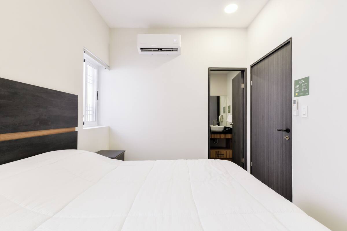 A bedroom with a bed, a nightstand, an air conditioning unit, a wardrobe, and a bathroom visible through an open door.