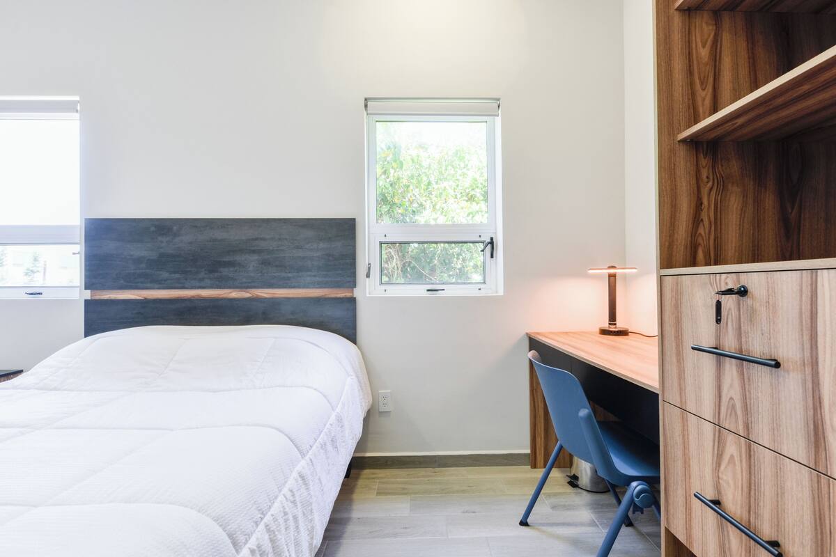 A bedroom with a bed, a desk, a chair, a wardrobe, and a window with a view of greenery.