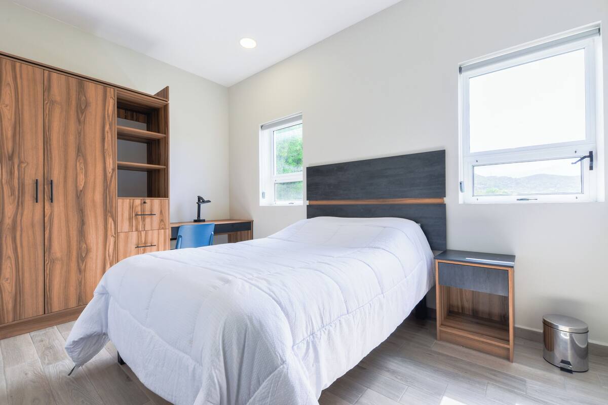 A bedroom with a bed, a wardrobe, a desk, a chair, a window, and a trash bin.