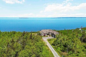 A large house with a driveway, surrounded by dense green trees and a view of a large body of water. - Fundy Bay Coastal Estates – Annapolis Valley (Victoria Harbour)