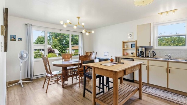 A modern kitchen with wooden cabinets, a dining area with a round table and chairs, a built-in oven, and a large window offering a view of greenery. - L`Acadie House in the Annapolis Valley (New Minas)