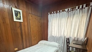 Un dormitorio con cama, escritorio con computadora portátil, silla, paredes de madera, un cuadro en la pared y una cortina.