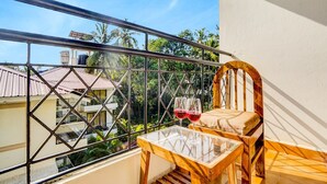 A balcony with a glass table and a wooden chair, two wine glasses on the table, and a view of buildings and trees. - Classic With Balconytownhouse Calangute (Calangute)