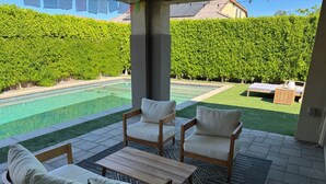 Eine Terrasse mit zwei Holzstühlen und einem Tisch, einem Pool und einer Hecke im Hintergrund.
