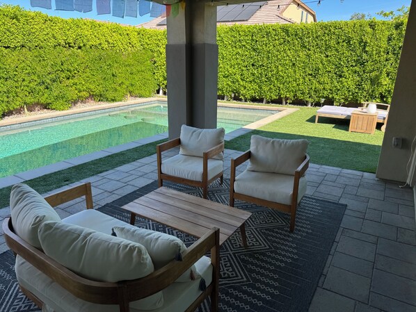 A patio with two wooden chairs and a table, a pool, and a hedge in the background. - Serene Oasis in the Desert- Trilogy at the Polo Club (Indio)