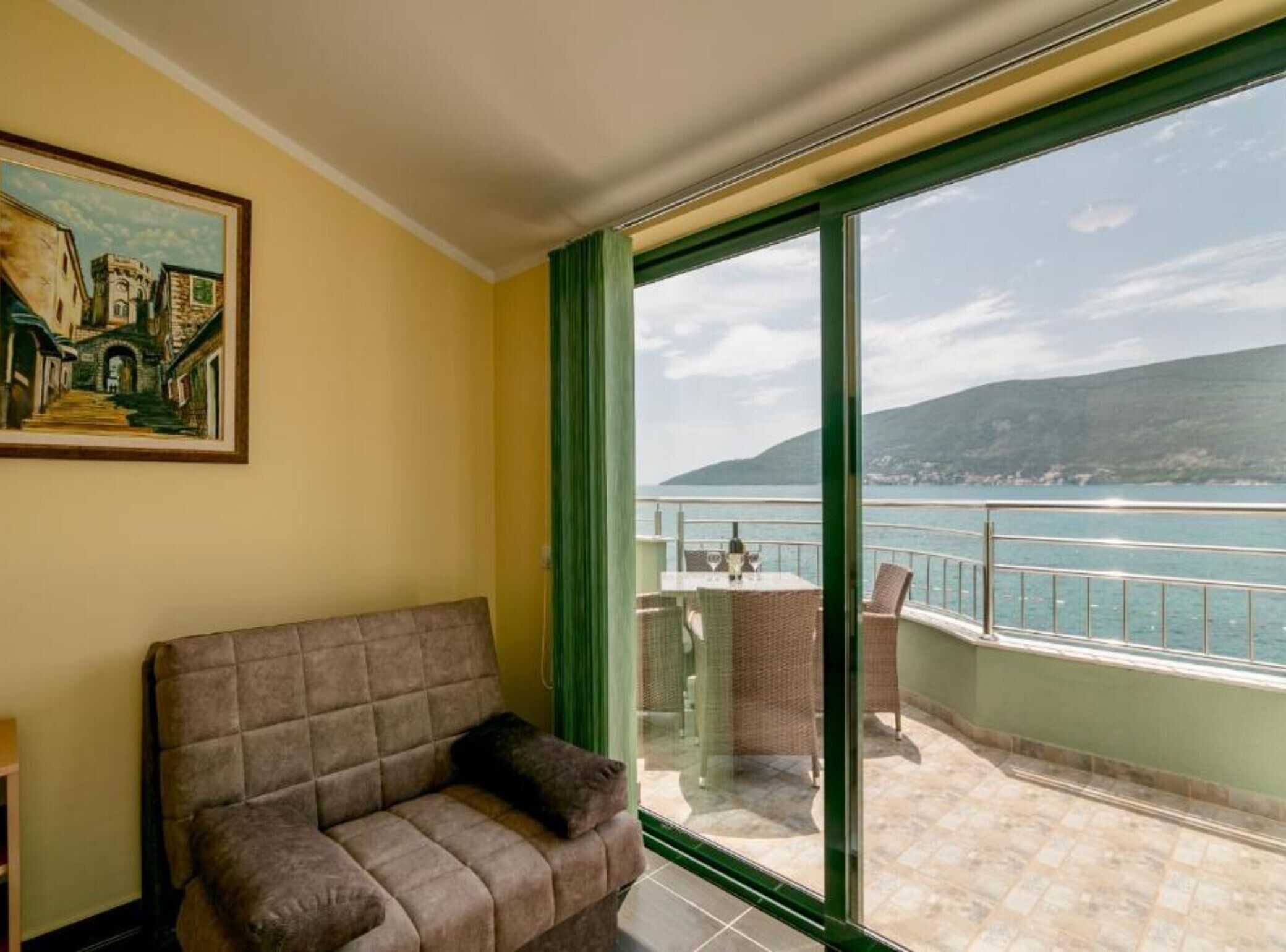 Une pièce comprenant un canapé, un tableau et un balcon avec une table et des chaises, d’où l’on peut admirer un plan d’eau et des collines.