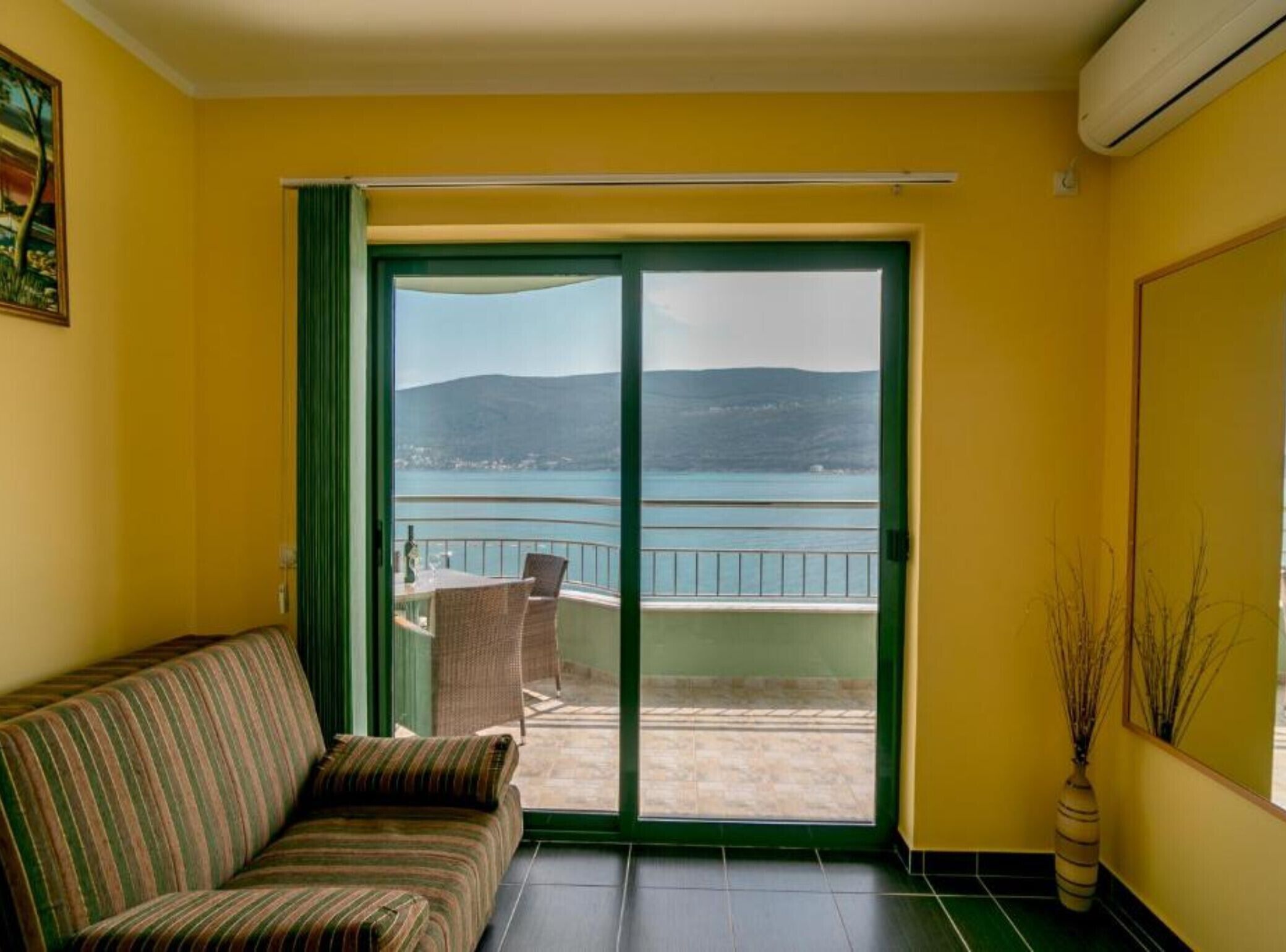 Een kamer met een bank, een schuifdeur naar een balkon met uitzicht op zee en bergen.