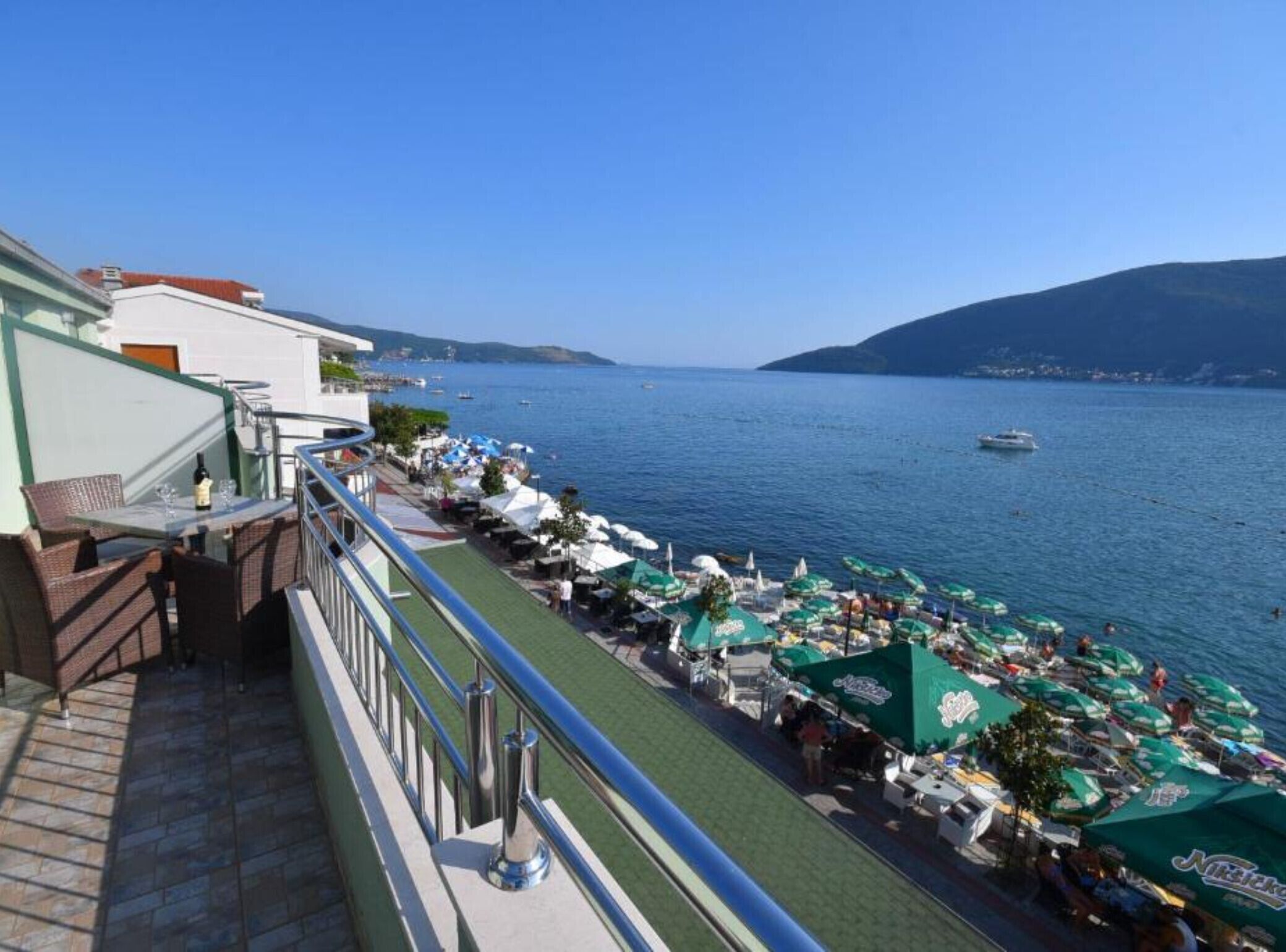 A balcony with outdoor seating overlooking a body of water, a distant boat, and a hilly landscape.