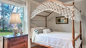 Een hemelbed met een witte hemel, een houten dressoir met lades, een lamp en een lijst met een afbeelding aan de muur.