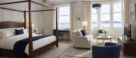 Una cama con dosel y vistas al mar, un rincón de descanso con una silla y una mesa, y un televisor sobre un mueble.