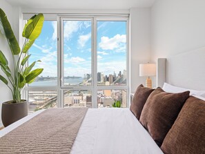 A hotel room with a large window, a bed, a plant, and a lamp.