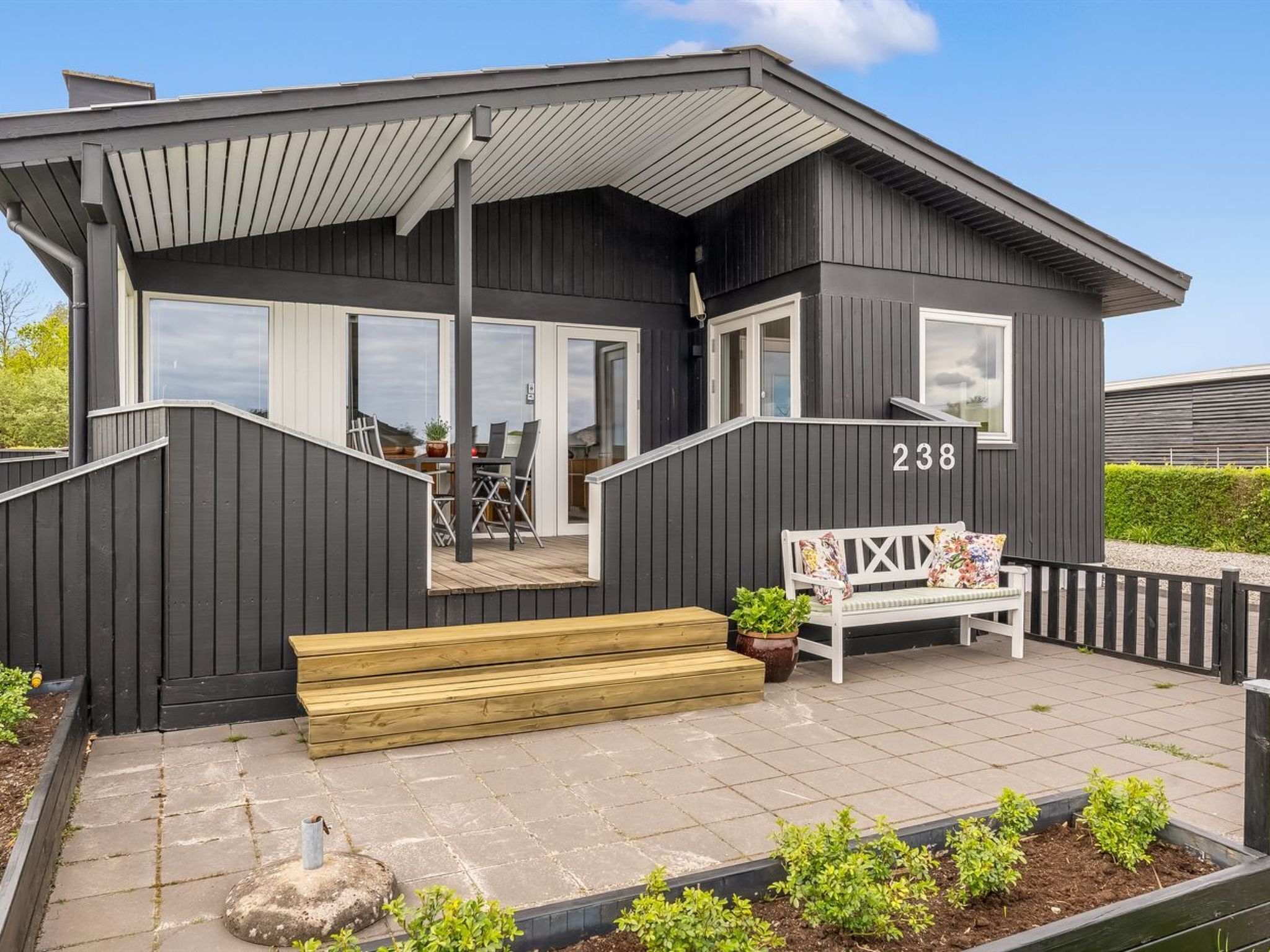 Ein modernes Haus mit der Nummer 238, einer überdachten Terrasse mit einer Bank und einem kleinen Garten.