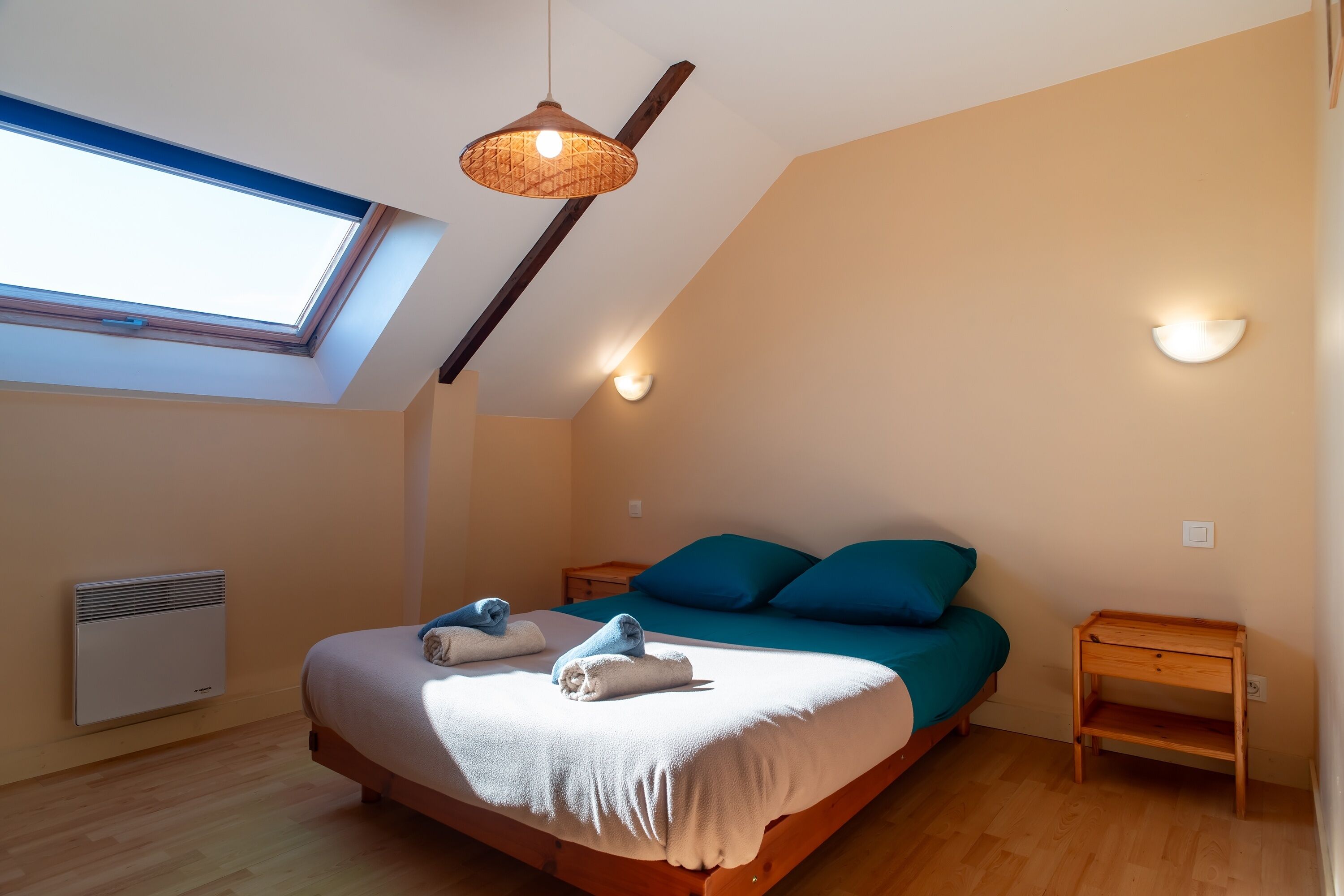 A bedroom with a skylight, a bed with blue bedding, a wooden nightstand, and a wall-mounted light fixture.
