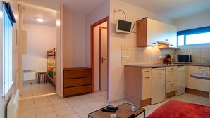 Una cucina compatta con forno a microonde, fornello e pensili, una piccola zona pranzo con tavolo e sedie, e un letto rosso in camera da letto.