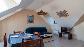 A dining area with a table set for two, a blue sofa, a television on a stand, and a framed picture on the wall. - Les Mésanges Bleues - Au Soleil Breton (Saint-Nic)
