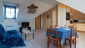 A cozy living room with a blue sofa, a dining table with chairs, a TV, and a kitchen area with a microwave. - Les Mésanges Bleues - Au Soleil Breton (Saint-Nic)