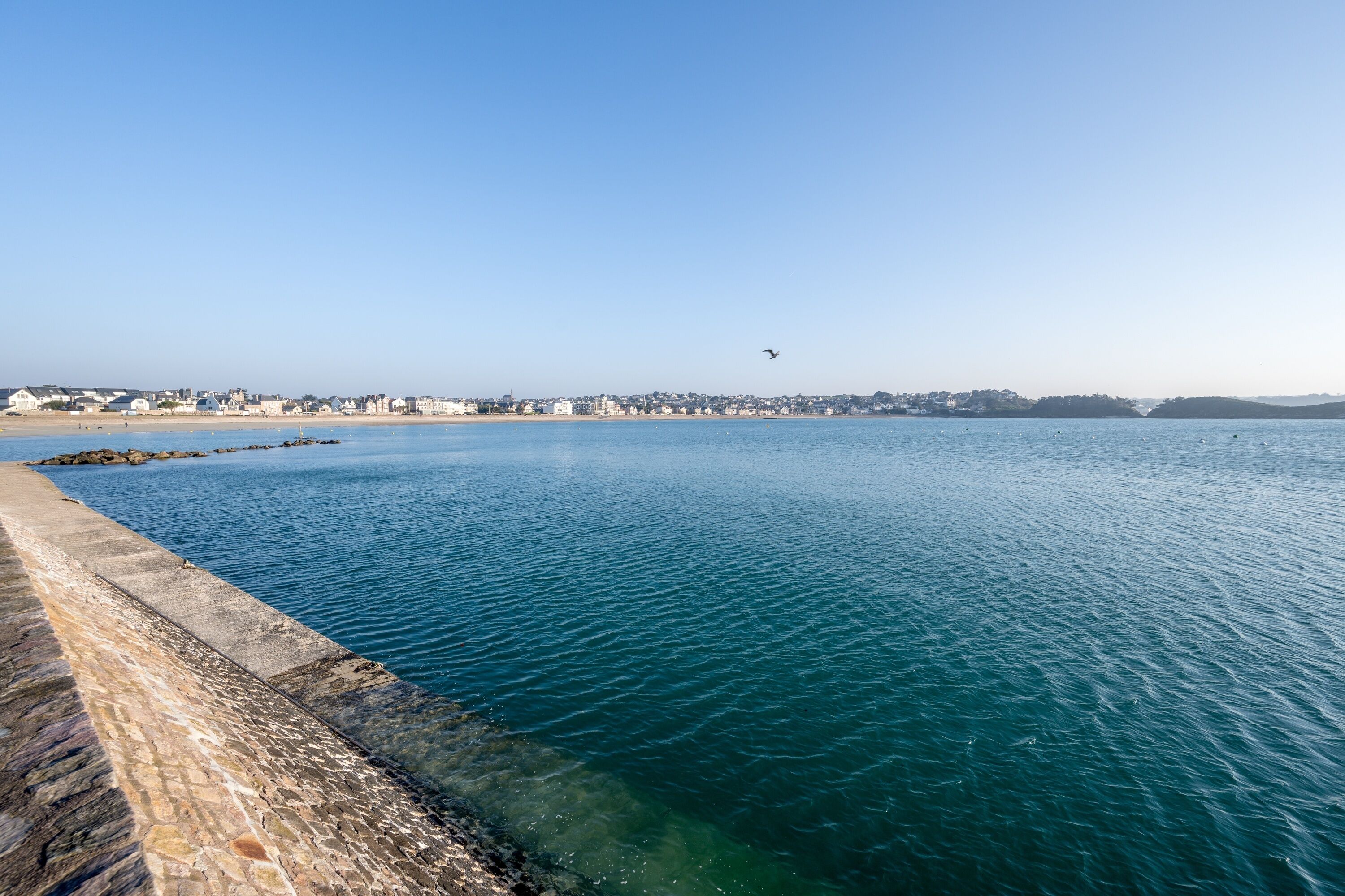 A coastal view with a clear sky, calm sea, and a stone pier extending into the water.