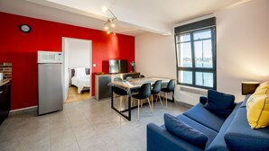 A modern apartment with a red accent wall, a dining area, a kitchenette, and a large window offering a view.