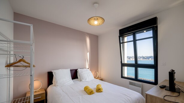 A hotel room with a bed, a bedside table, a desk, a chair, a window with a view, and a hanging light fixture.