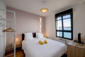 Habitación de hotel con cama, mesita de noche, escritorio, silla, ventana con vistas y lámpara colgante.