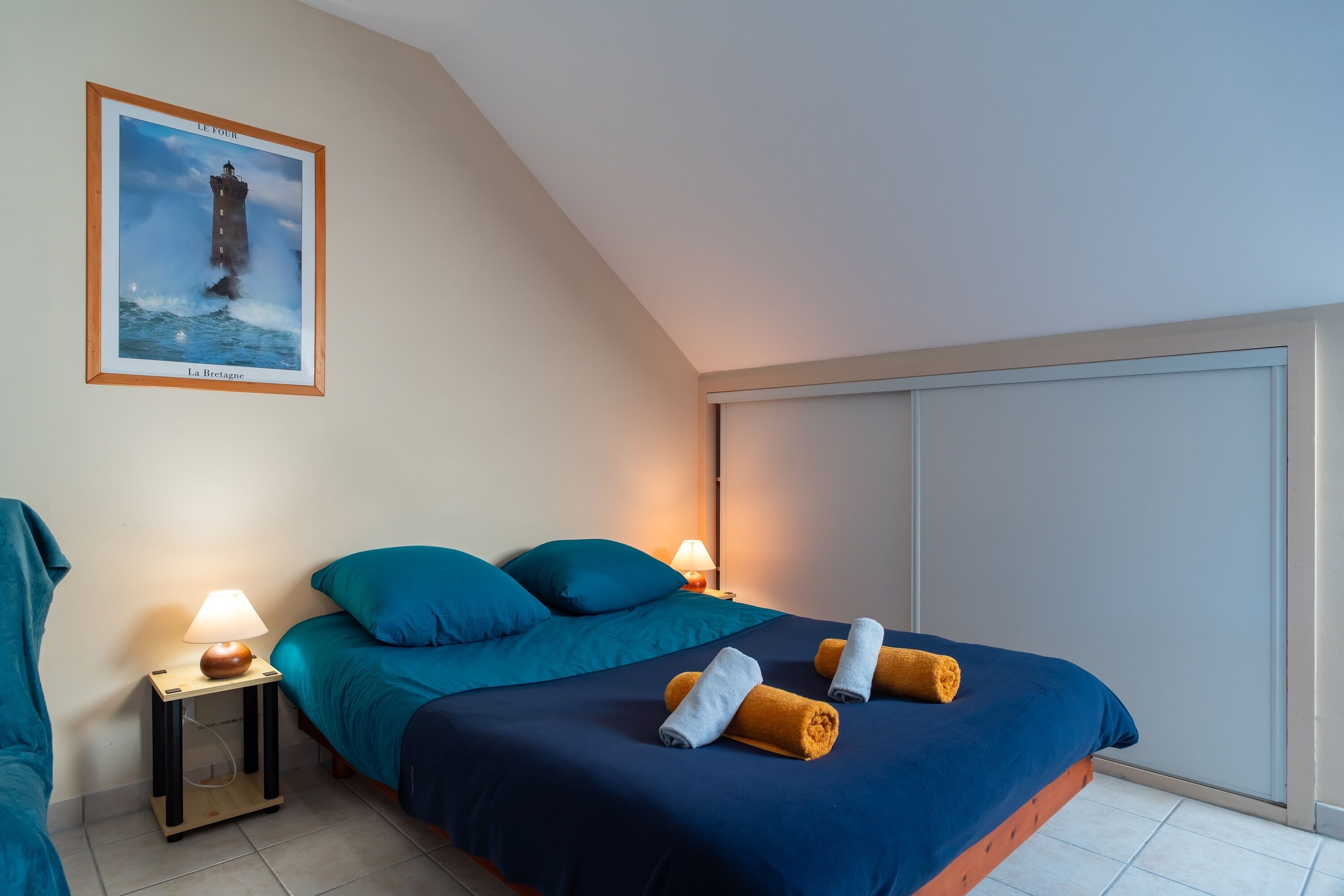 A bedroom with a bed, two bedside tables with lamps, a framed picture on the wall, and a large window.