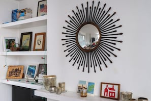 A room with a sunburst mirror, shelves with books and framed pictures, and decorative items on a mantel. - Artistic 1BDR Townhome | Central Brighton (Brighton and Hove)