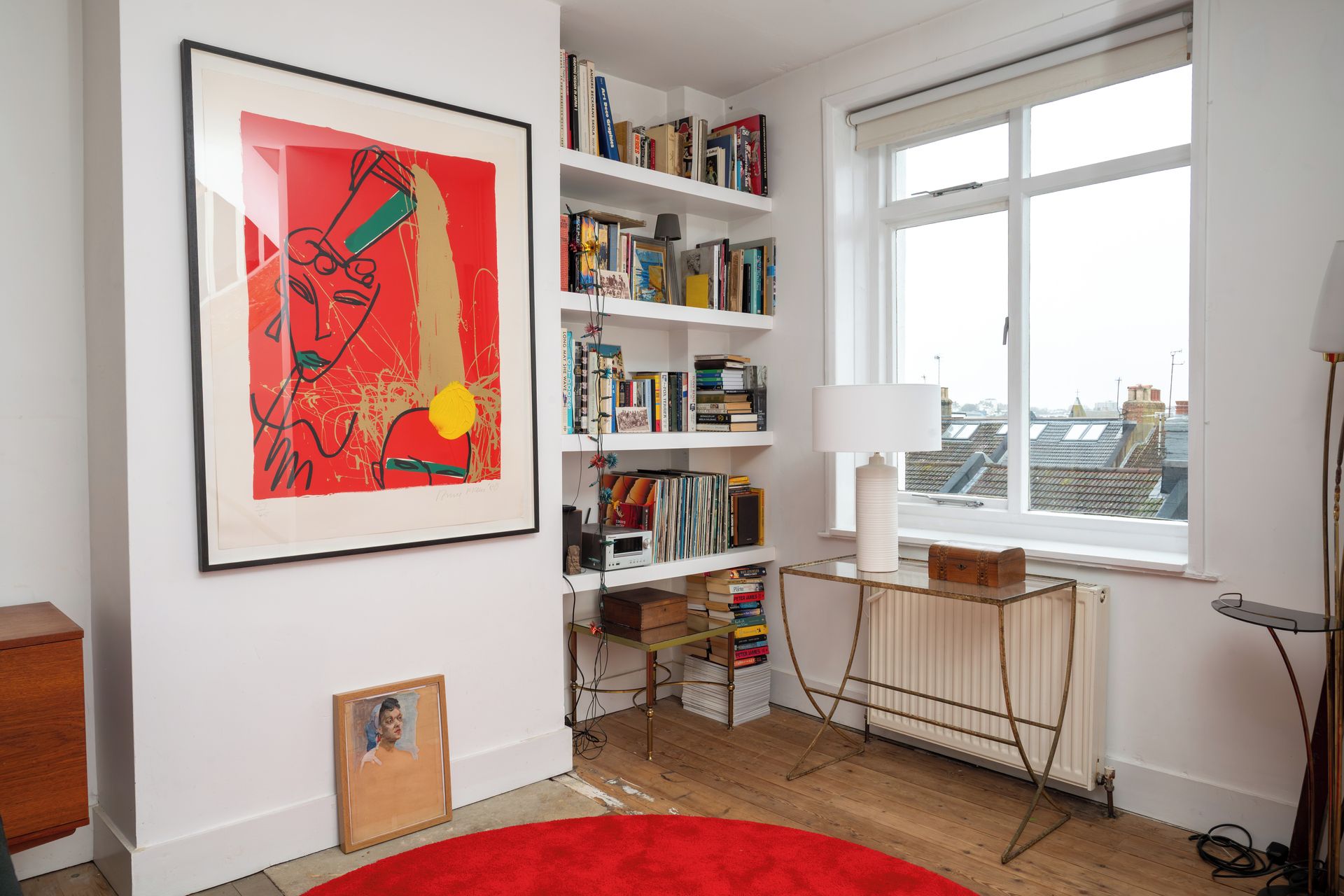 Een kamer met een groot abstract schilderij, een boekenplank vol boeken, een radiator en een raam met uitzicht op gebouwen.