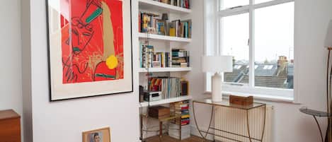 A room with a large abstract painting, a bookshelf filled with books, a radiator, and a window with a view of buildings.