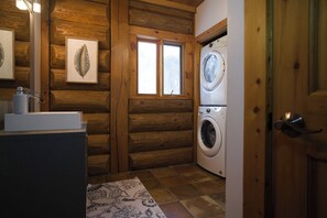 Une pièce aménagée dans le style d’un chalet en rondins, avec un lave-linge, un évier avec un miroir et une fenêtre décorée d’un motif représentant des feuilles.